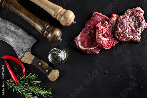 Piece of beef tenderloin, with ax for cutting and chopping meat, spices were cooking - rosemary, pepper on old black table. Top view copy space photo