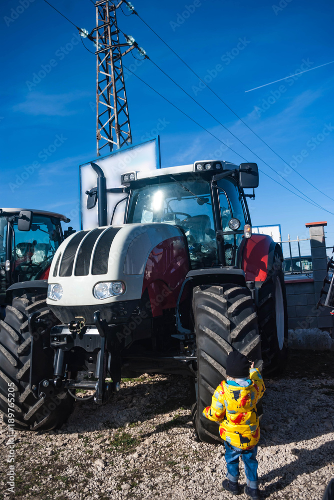 Agricultural machinery. Agro-technology. Front view of the new shiny tractor and littele boy near the rubber. Agricultural machinery. Agro-technology. Front view of the new shiny tractor and littele boy near the rubber.
