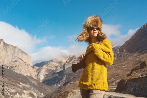 A girl photographer in sunglasses and a big fur hat and a yellow knitted sweater stands against the background of high rocks in the gorge with a dslr camera her in hand.