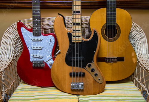 Group of musical instruments: electric bass, red electric guitar and classic or spanish guitar on wicker armchair. Indoor place.