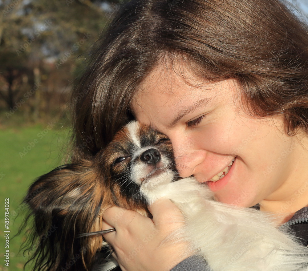 A young girl with long brown hair holds a little hairy dog - her pet. She is happy and smiles and the dog looks like smart. They are in the garden or park with grass. It is a concept of friendship. A young girl with long brown hair holds a little hairy dog - her pet. She is happy and smiles and the dog looks like smart. They are in the garden or park with grass. It is a concept of friendship.