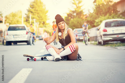 Cool skater girl in street style, sitting on the street posing with skate board in urban environment