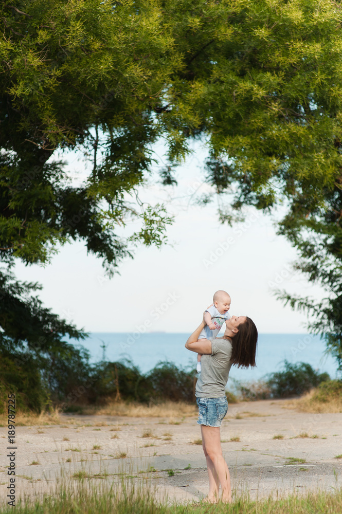 Young fun smiling woman standing at sea background hugging little cute child baby boy on nature, green trees. Mother, little kid son. Parenthood, family day 15 of may, love, parents, children concept.