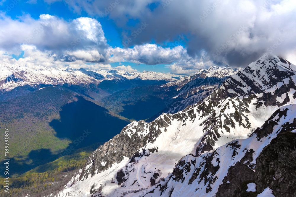 Fototapeta premium Beautiful panoramic view of Aibga Ridge from the top of the mountain at a height of 2320 meters with huge white clouds casting a shadow. Ski Resort at Caucasus Mountains, Rosa Peak, Sochi, Russia.