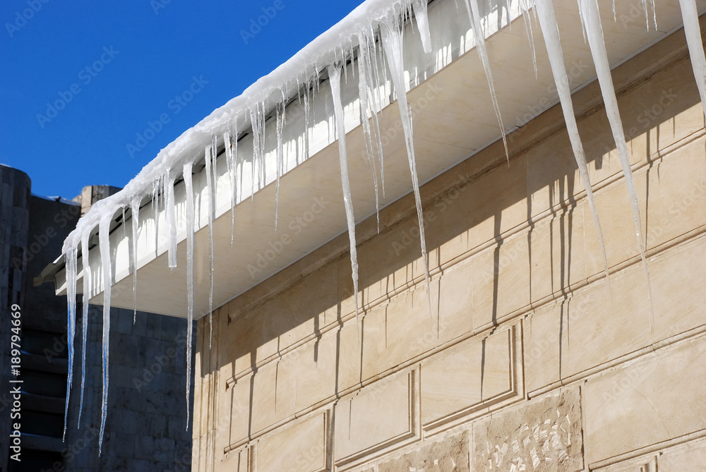 Fototapeta premium Winter. Huge dangerous ice icicles hang above the street threat health and life of people hanging from the roof of building with cracked walls. Frozen air conditioner with huge piece of ice pendant