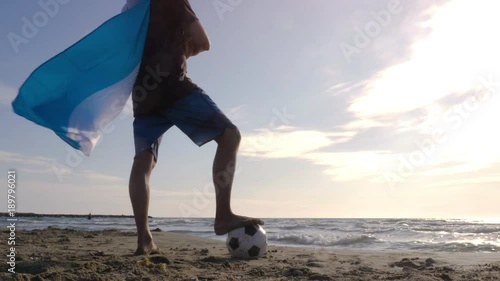 Young man wearing argentinian argentina national flag as super hero cape stands with foot on football on the sea shore sand looking at the ocean at the beach at sunset camera steadycam gimbal photo