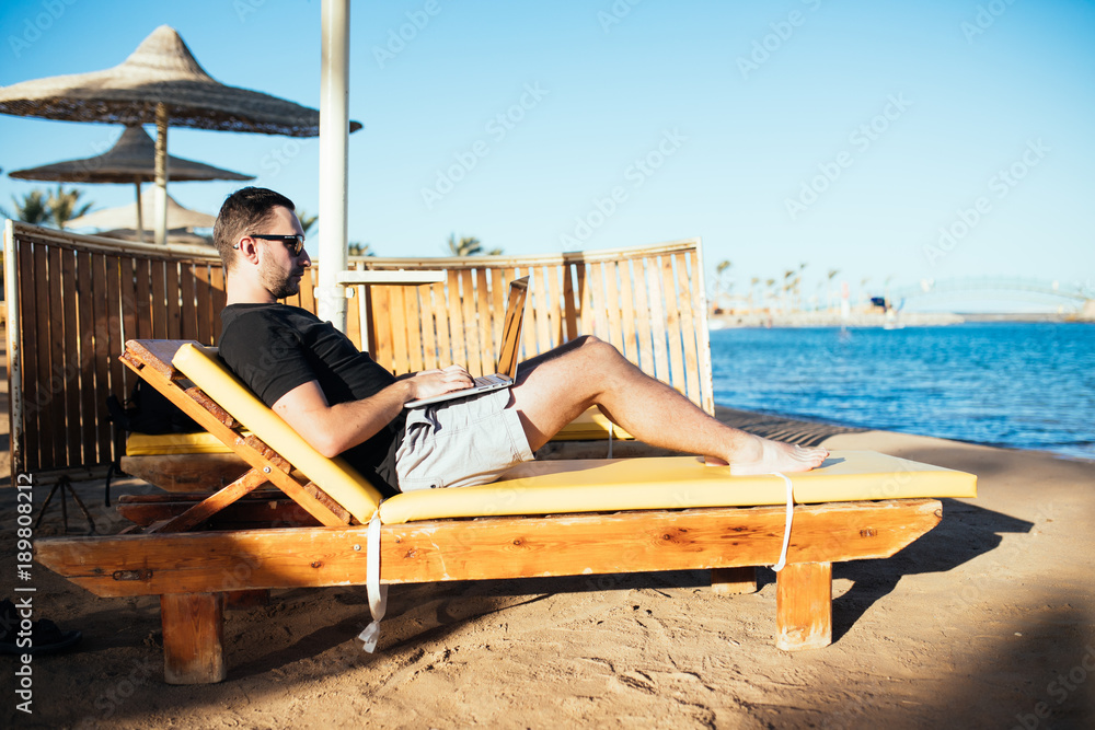 Young handsome businessman in sunglasses working with laptop lying on the deck at the beach sea