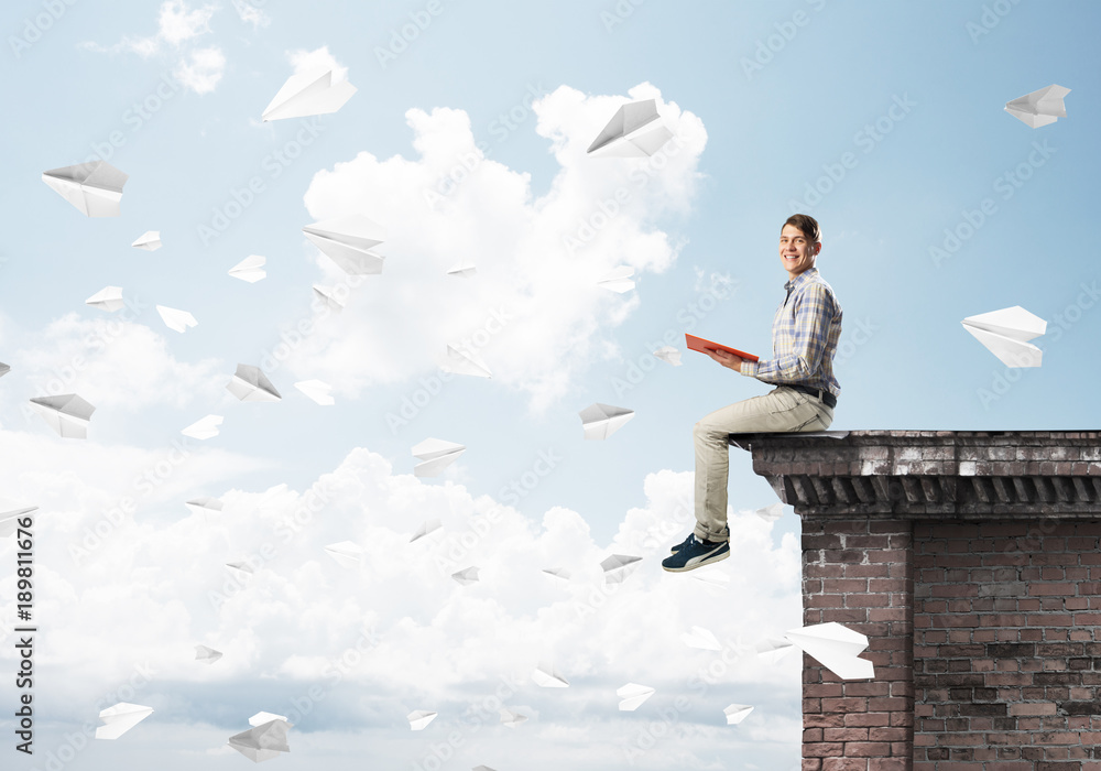 Handsome student guy on roof edge reading book and paper planes fly around Handsome student guy on roof edge reading book and paper planes fly around