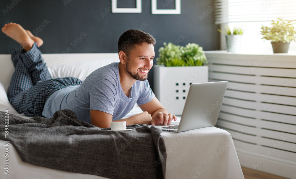 happy young man with laptop and cup of coffee in bed