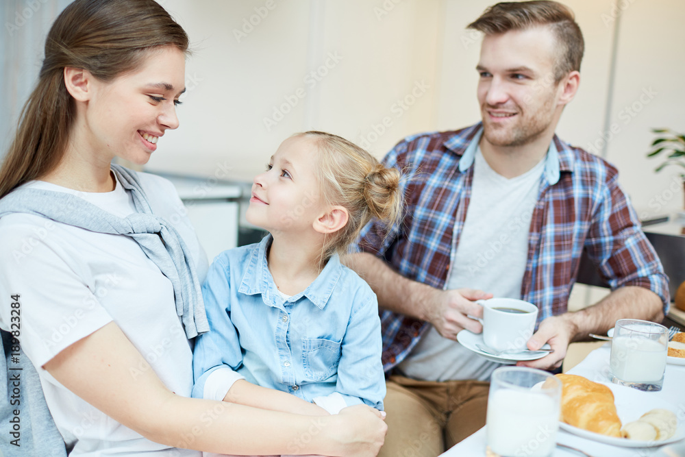 Sweet little girl looking at her mother during breakfast, father sitting near by and listening to them Sweet little girl looking at her mother during breakfast, father sitting near by and listening to them