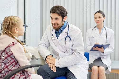 Smiling young clinician listening to little patient complaints while his assistant making notes