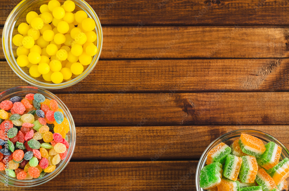 Brightly colored candies in glass cups on a wooden table, top view, free space for text Brightly colored candies in glass cups on a wooden table, top view, free space for text