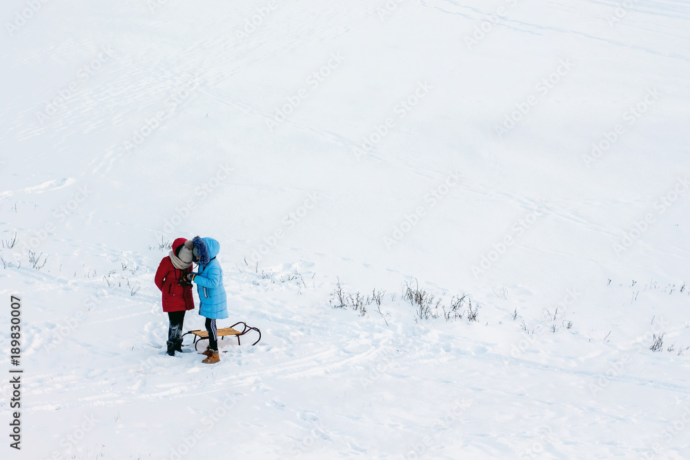 two girls with sleds in an open area covered with snow, something is being considered