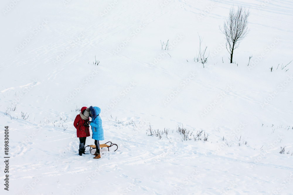 two girls with sleds in an open area covered with snow, something is being considered