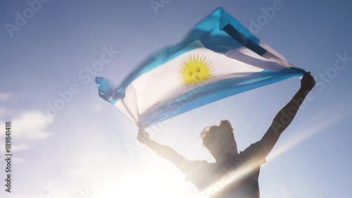 Young man holding argentinian national flag to the sky with two hands at the beach at sunset argentina
