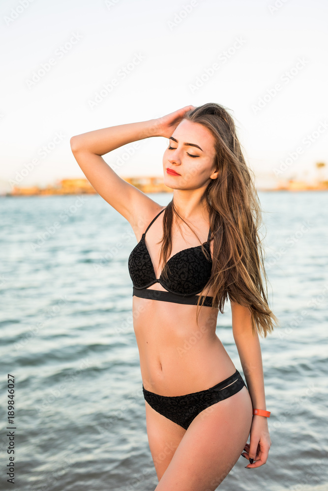 Young beautiful girl in white swimsuit bikini posing on beach in sunlight. Sunny portrait.