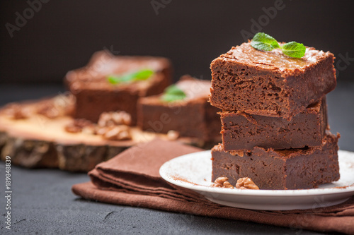 Chocolate brownie square pieces in stack on white plate with walnuts, decorated with mint leaves and cocoa on black background. Delicious dessert. Dark mood. Close up photography. Selective focus