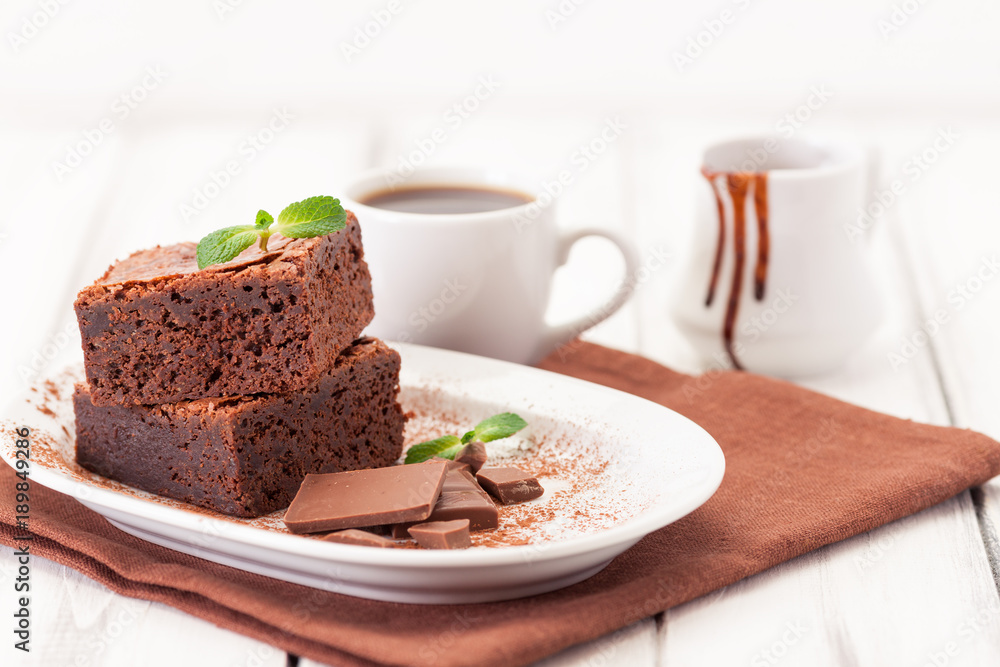 Chocolate brownie square pieces in stack on white plate decorated with mint leaves and cocoa powder on white vintage wooden background. American traditional delicious dessert. Close up photography Chocolate brownie square pieces in stack on white plate decorated with mint leaves and cocoa powder on white vintage wooden background. American traditional delicious dessert. Close up photography