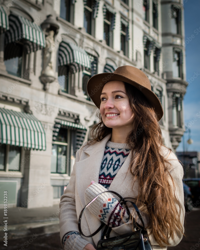 Portrait of a young female tourist with elegant clothes exploring the city. Trendy woman traveler with smile looking on buildings and touring old town. Beautiful girl walks in Rotterdam, Netherlands.