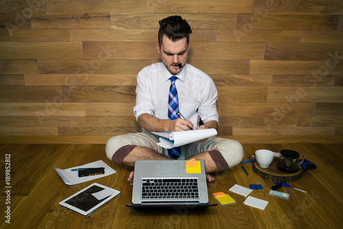 A barefoot businessman sits on the floor against a wall with a lot of gadgets around him. Serious young man works at home in funny clothes and makes important notes