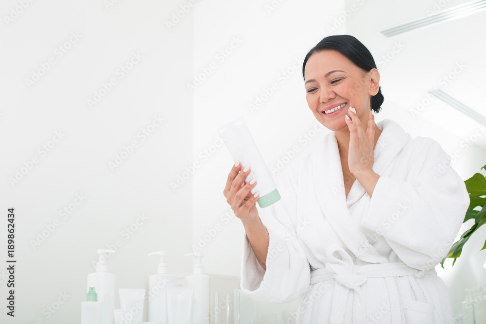 Glad asian lady reading the instruction in beauty salon. She is looking at tube of cream in her hand while touching skin. Copy space in left side Glad asian lady reading the instruction in beauty salon. She is looking at tube of cream in her hand while touching skin. Copy space in left side