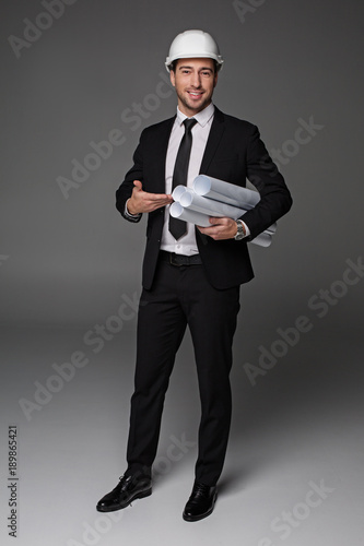 Full length portrait of beaming bearded master-builder pointing at plans by arm. Labor concept