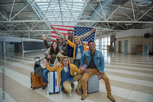 Real patriots. Full length portrait of joyful young friends are posing at airport lounge with usa flag. They are sitting on suitcases while looking at camera with joy and expressing happiness