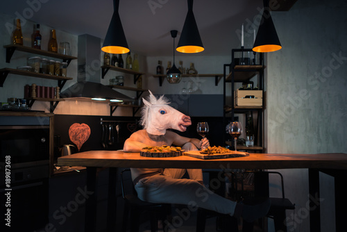 Young man sitting at the table with food and drinking wine in modern home kitchen. Funny unicorn dinning alone at the bar counter in stylish apartment. Crazy person. Painted heart on the wall
