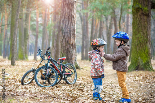 Two brothers preparing for bicycle riding in spring or autumn forest park. Older kid helping sibling to wear helmet. Safety and protection concept. Happy boys best friends having good time together.