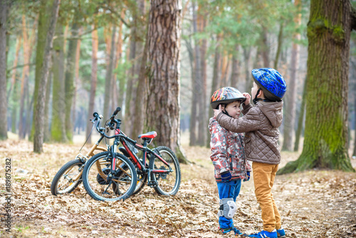 Two brothers preparing for bicycle riding in spring or autumn forest park. Older kid helping sibling to wear helmet. Safety and protection concept. Happy boys best friends having good time together.