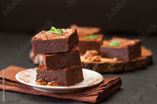 Chocolate brownie square pieces in stack on white plate with walnuts, decorated with mint leaves and cocoa on black background. Delicious dessert. Dark mood. Close up photography. Selective focus