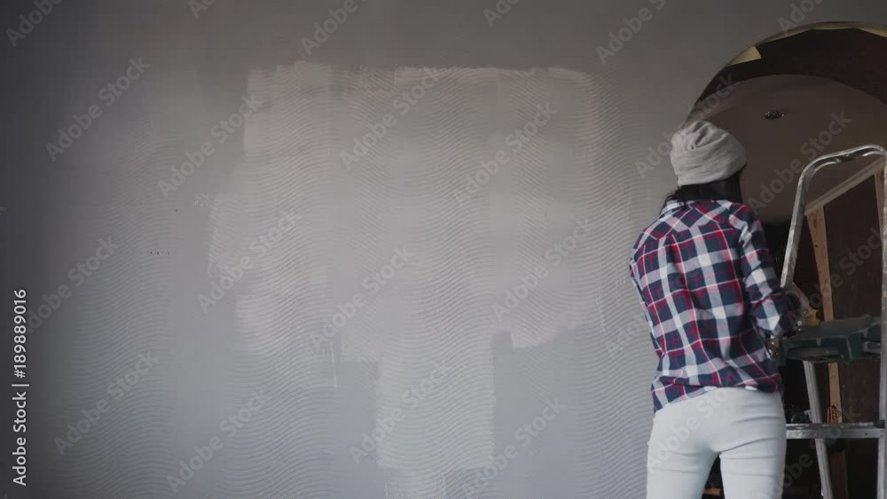 A young woman in a gray hat draws a wall in her new home at gray color, listens to music from the phone in headphones and fun dancing A young woman in a gray hat draws a wall in her new home at gray color, listens to music from the phone in headphones and fun dancing