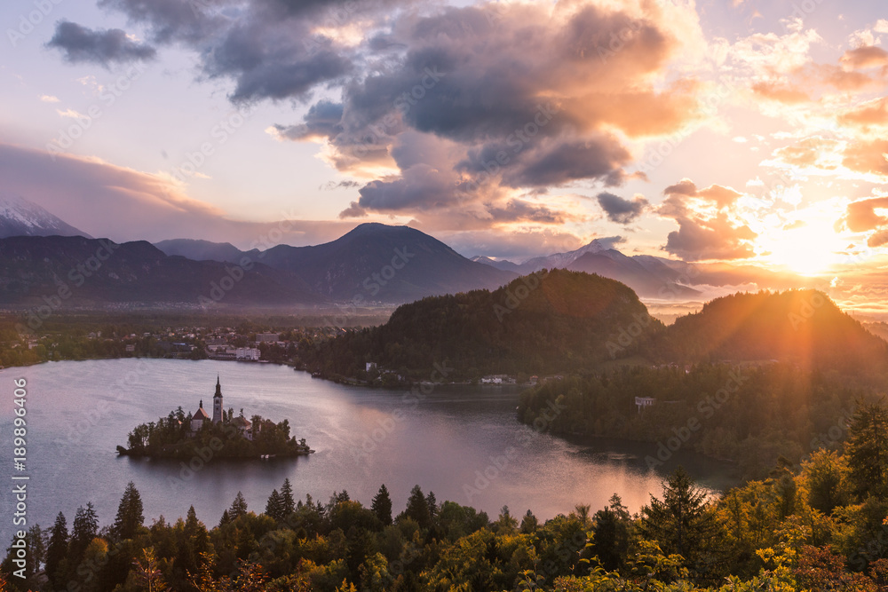 Sun peaking through clouds looking at Lake Bled in Slovenia. The sunrise shows purples and oranges from the classic viewpoint. A layer of fog is in the distance near the town.