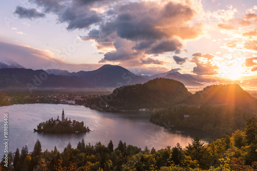 Fototapeta Naklejka Na Ścianę i Meble -  Sun peaking through clouds looking at Lake Bled in Slovenia. The sunrise shows purples and oranges from the classic viewpoint. A layer of fog is in the distance near the town.