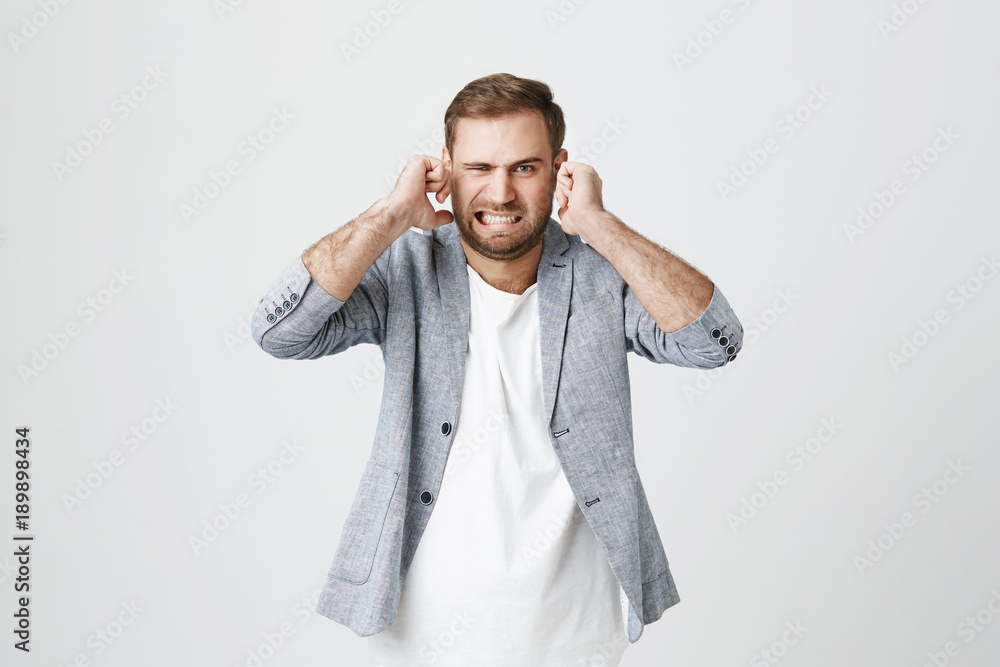 Irritated handsome bearded male with displeased facie expression, plugs ears, clenches teeth, expresses negativeness, avoids noise, isolated against gray background. Stop this annoying sound! Irritated handsome bearded male with displeased facie expression, plugs ears, clenches teeth, expresses negativeness, avoids noise, isolated against gray background. Stop this annoying sound!