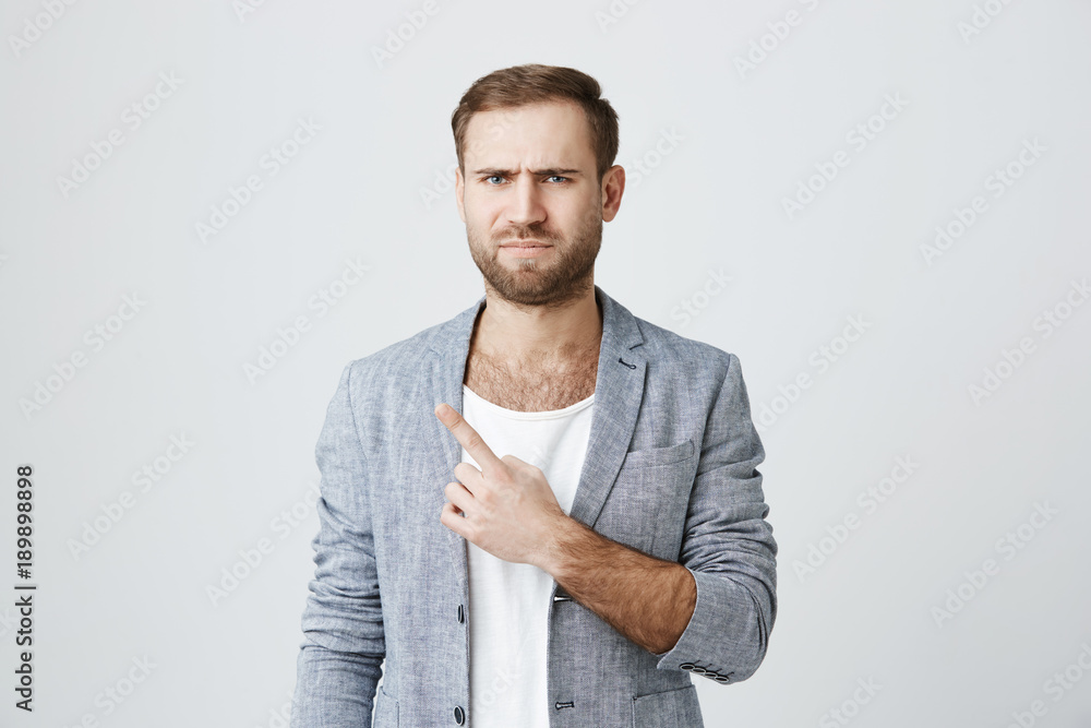 Negative emotions and feelings. Dissatisfied european man with stubble frowning face looking angrily at camera, pointing with finger at copy space on blank studio wall. Negative emotions and feelings. Dissatisfied european man with stubble frowning face looking angrily at camera, pointing with finger at copy space on blank studio wall.
