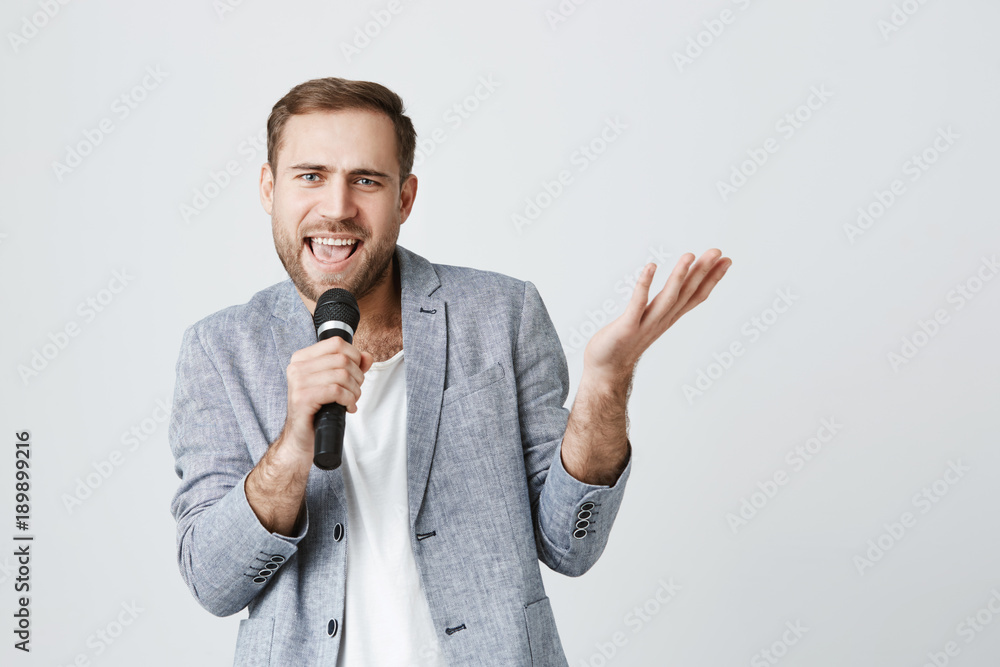 Talented bearded singer in trendy gray jacket holding microphone singing his best song. Office worker spends evening by singing at bar, having fun. Music and leisure time concept. Talented bearded singer in trendy gray jacket holding microphone singing his best song. Office worker spends evening by singing at bar, having fun. Music and leisure time concept.