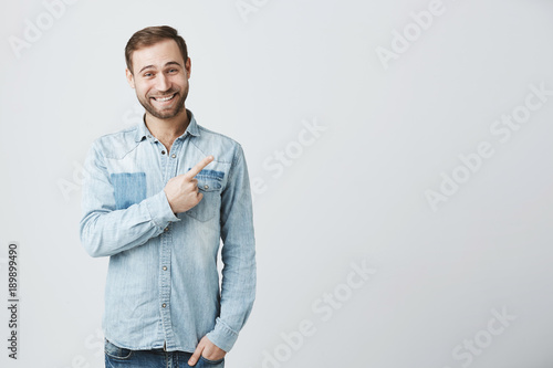 Caucasian young male in denim shirt grins at camera, indicates at copy space, advertises something. Happy bearded man points with fore finger, has satisfied expression, smiles. Look here!
