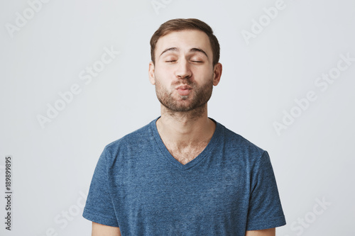 Attractive good-looking bearded male model closes eyes, pouts lips, sends kisses to camera, poses against gray background. Handsome european man with appealing appearance