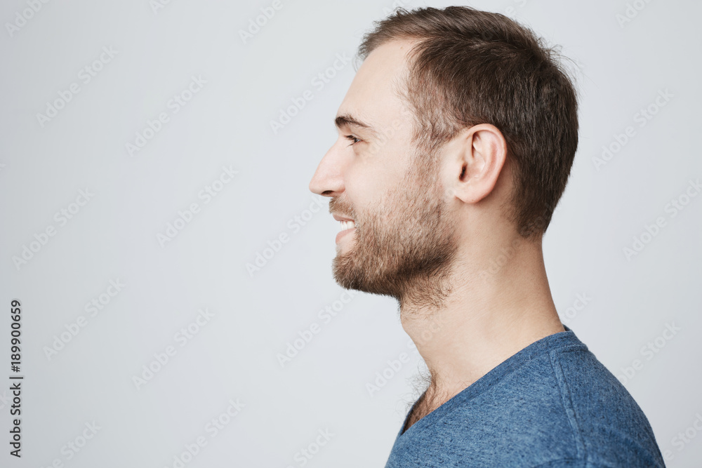 Obraz premium Close up profile of stylish smiling attractive male with stubble and dark hair wearing blue t-shirt posing against gray blank wall with copy space for your text or advertising content