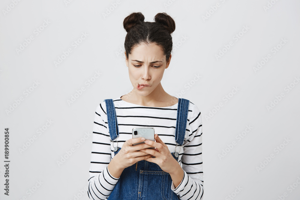 Shocked brunette female with hairbuns holds mobile phone, missed important call, being puzzled. Horrified girl bites lower lip looks at watch on smart phone, realizes that she is late on meeting Shocked brunette female with hairbuns holds mobile phone, missed important call, being puzzled. Horrified girl bites lower lip looks at watch on smart phone, realizes that she is late on meeting
