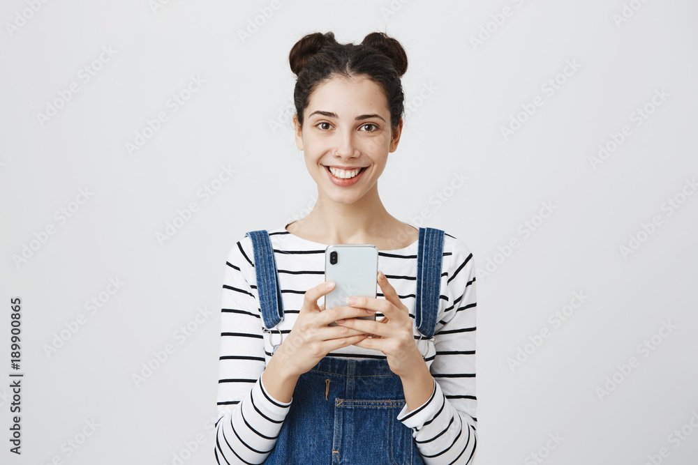 Happy brunette young woman with hair knots, wears denim overall, uses smartphone for taking pictures, smiles pleasantly, isolated against gray background. People and happiness concept