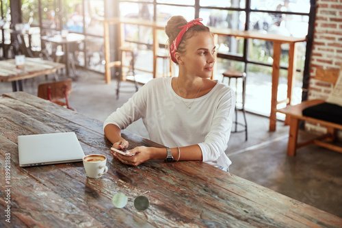Beautiful woman wears casual white sweater and red bandage views photos in social networks, chats with friends online while sits in coffee bar. Creative copywriter searches information on cellular