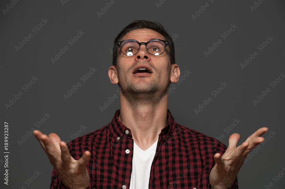 Naklejka premium Portrait of a man in casual clothes and glasses looks up enthusiastically and extends his palms against gray background