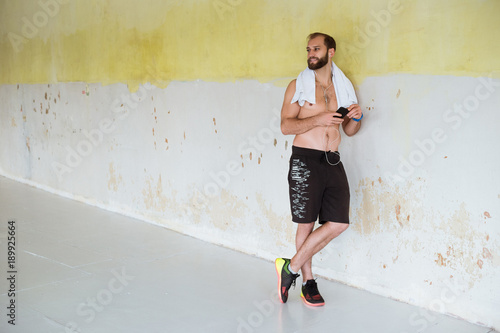 A young happy sports man staying near the wall in the gym and holding the phone.