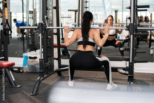 Back view of beautiful young woman training with barbell in modern gym standing against window, copy space