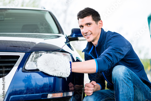 Smiling man cleaning the headlamp on his car wiping it with a mitt as he crouches alongside the vehicle
