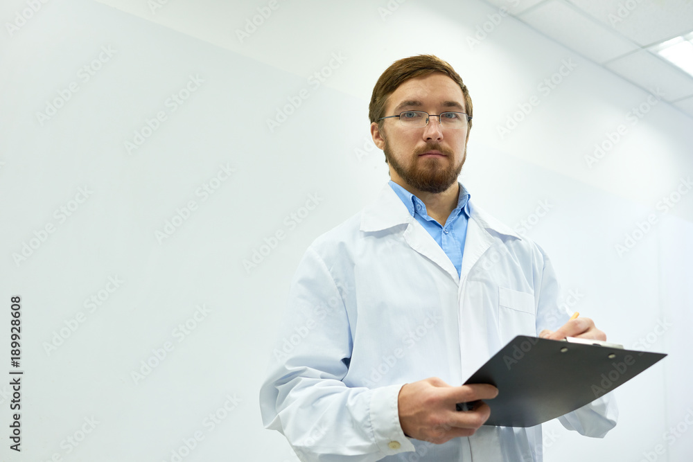 Low angle portrait of bearded doctor wearing glasses standing against white wall writing on clipboard and looking at camera, copy space Low angle portrait of bearded doctor wearing glasses standing against white wall writing on clipboard and looking at camera, copy space
