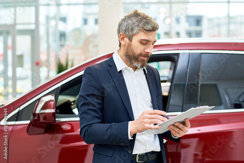 Wallpaper Mural Portrait of handsome mature salesman making notes on clipboard  standing by brand new luxury car in showroom, copy space Torontodigital.ca