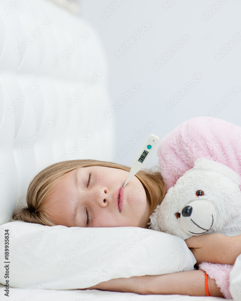 sick girl with toy bear lying in bed with a thermometer in mouth
